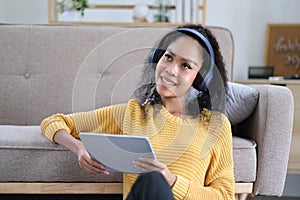 Photo of beautiful dark skin wavy lady homey mood holding digital tablet earflaps listening new audio sitting floor near couch