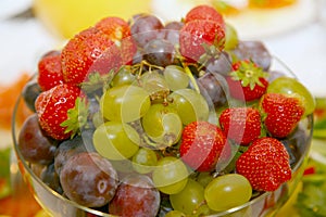 Photo of beautiful and delicious fruits and berries, grapes, strawberries and plums lying heaped in a round glass vase.