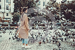 Photo of beautiful girl looking at pigeons on the square, flocking wings in cloudy autumn day.