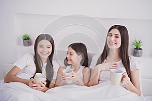 Photo of beautiful pretty three sisters sleepwear sitting bed having rest laughing drinking morning beverages inside