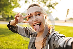 Photo of beautiful sporty woman 20s in sportswear wearing bluetooth earpod taking selfie with fingers, while walking in green park