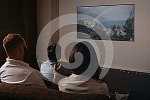 A photo from behind of a family which is watching a movie on a widescreen TV set