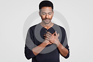 Photo of black man with faithful expression, keeps hands on chest, expresses sympathy, closes eyes, feels gratitude, dressed in