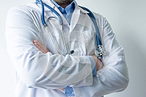 Photo of body of doctor in white lab coat with stethoscope on his neck in half-turn with folded hands against white wall in hospit