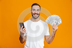 Photo of brunette man 30s in casual wear holding cell phone and fan of money, isolated over yellow background