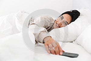 Photo of brunette woman 30s using smartphone, while lying in bed with white linen at home