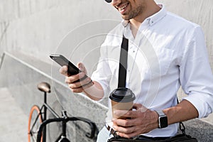 Photo of businesslike man 30s wearing sunglasses, drinking takeaway coffee and using mobile phone while standing with bicycle