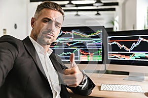 Photo of businesslike man taking selfie while working in office on computer with graphics and charts