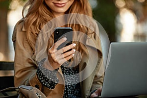 Photo of businesswoman using mobile phone and laptop while sitting on bench in sunlit alley