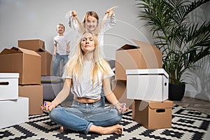 Photo of calm blonde sitting on floor among cardboard boxes and boy, girl jumping on sofa