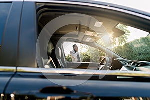 Photo of the car window on sunny day and successful African businessman walking while taking at the phone on the