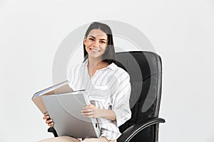 Photo of caucasian brunette businesswoman 30s smiling and holding bunch of paper folders while sitting in black armchair in office