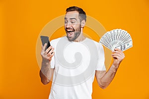 Photo of caucasian man 30s in casual wear holding cell phone and fan of money, isolated over yellow background