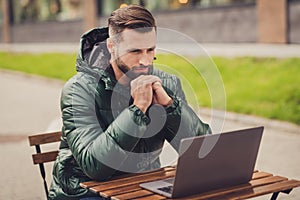 Photo of charming attentive young man wear green windbreaker reading modern device having rest cafe outside city street