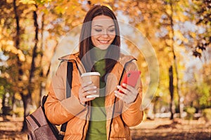 Photo of charming lady smiling telephone look screen cup drink takeout coffee greeting friend online autumn park wear