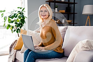 Photo of charming lovely retired lady sittin sofa using modern netbook chatting with customers clients online at home