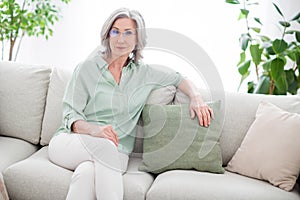 Photo of charming serious calm old woman sit sofa weekend wear glasses indoors inside house home flat