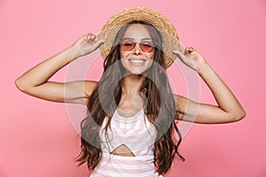 Photo of charming woman 20s wearing sunglasses and straw hat smiling at camera, isolated over pink background