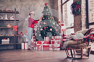 Photo of charming young girl hang balls x-mas tree wear santa headwear red sweater decorated living room indoors