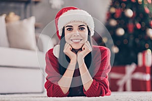 Photo of charming young girl lay carpet fists chin toothy smiling wear santa headwear red pullover in decorated living