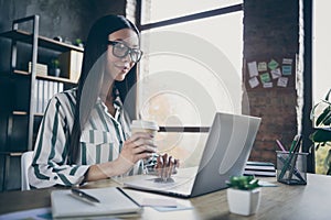 Photo of cheerfu smiling happy joyful girl holding disposable cup of coffee sitting at table with laptop on it looking