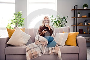 Photo of cheerful adorable cute love pensioner woman sitting on comfortable sofa turning on tv drinking tea indoors