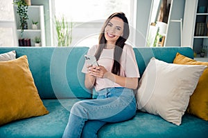 Photo of cheerful adorable girl toothy smile sit cozy couch hold telephone bright modern flat indoors