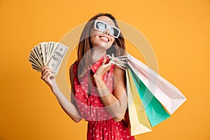 Photo of cheerful elegant young woman in sunglasses and red dress holding fan of money and colorful shopping bags, looking aside