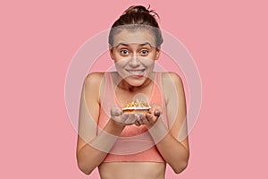 Photo of cheerful fitness lady feels temptation while holds delicious sweet cake, wears casual top, bites lips, doesnt