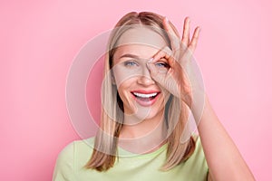 Photo of cheerful positive foolish lady make okey sign cover eye wear green t-shirt isolated on pink background