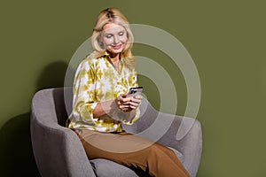 Photo of cheerful relaxed senior age woman sit on grey armchair she surfing news feed in smartphone isolated over khaki