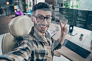 Photo of cheerful web designer guy sit chair take selfie demonstrate v-sign open space workstation indoors