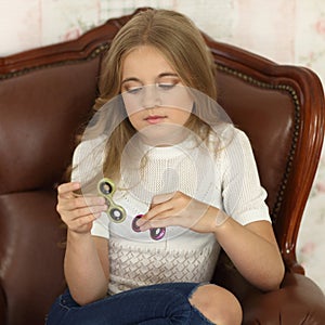 Photo of a child with green and pink fidget spinners in hands, sitting in leather chair, wearing white shirt and jeans.