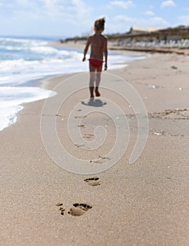 A photo with children`s footprints in the sand of the beach in the foreground and a little girl walking along the sea shore bac