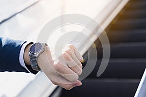 The photo close up of businessman watching his watch in his hand while standing on the escalator.  Business and finance concept