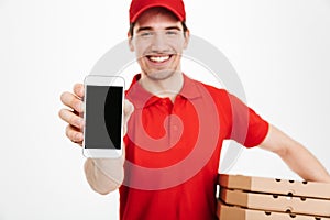 Photo closeup of affable man from delivery service in red t-shirt and cap holding stack of pizza boxes and showing