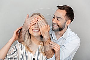 Photo closeup of handsome man in casual clothing smiling and covering eyes of his girlfriend