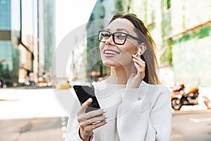 Photo closeup of happy smiling woman using cellphone and earpod while walking in big city street