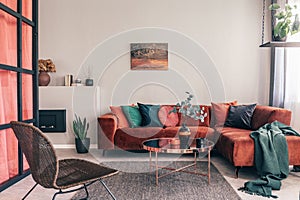 photo of a comfy living room interior with a round table on gray rug, wicker armchair and red corner sofa