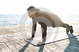 Photo of concentrated sportsman 20s in tracksuit lying on fitness mat and doing plank exercise by seaside