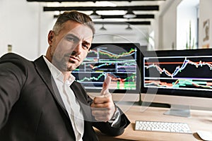 Photo of confident man taking selfie while working in office on computer with graphics and charts at screen