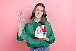 Photo of confident pretty woman wear green shirt holding little house showing okey sign isolated pink color background