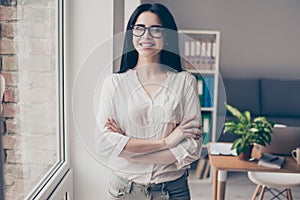 Photo of confident pretty young beautiful woman with spectacles stands edgeways in the office near the window, crossed