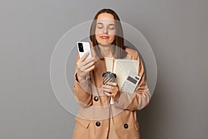 Photo of confused Caucasian woman wearing beige jacket holding organizer and calculator isolated over gray background, looking at