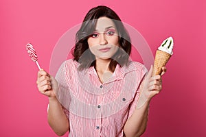 Photo of confused pensive sweet female holding ice cream and lollipop in both hands, looking directly at camera, having bright
