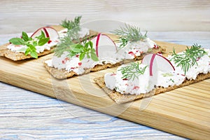 A photo of crackers, crisp rye bread toast with cottage cheese decorated with radish, cucumber, dill and basil leaves the wooden b