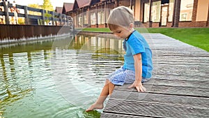 Photo of cute 3 years old little boy sitting on the riverebank at water canal in old town and splashing water with feet.