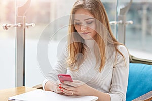 Photo of cute blonde female read something on her smart phone while sitting in coffee shop, beautiful young girl dressed casually photo
