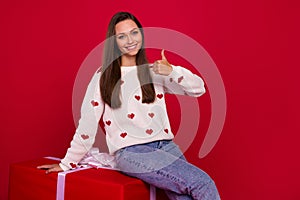 Photo of cute confident girl dressed white pullover showing thumb up sitting gift package isolated red color background