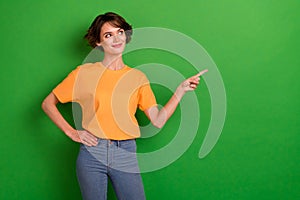 Photo of cute dreamy young woman dressed orange t-shirt pointing looking empty space isolated green color background